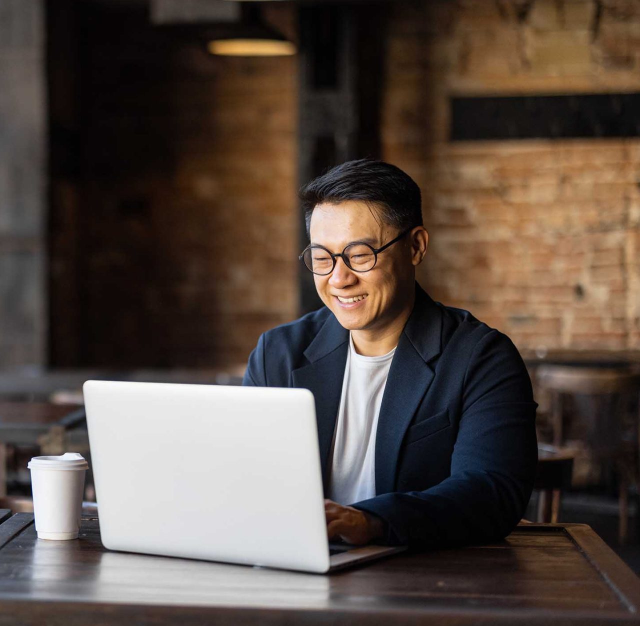 Asian businessman typing on laptop during work in cafe. Concept of remote and freelance work. Smiling adult successful man wearing suit and glasses sitting at wooden desk. Sunny day