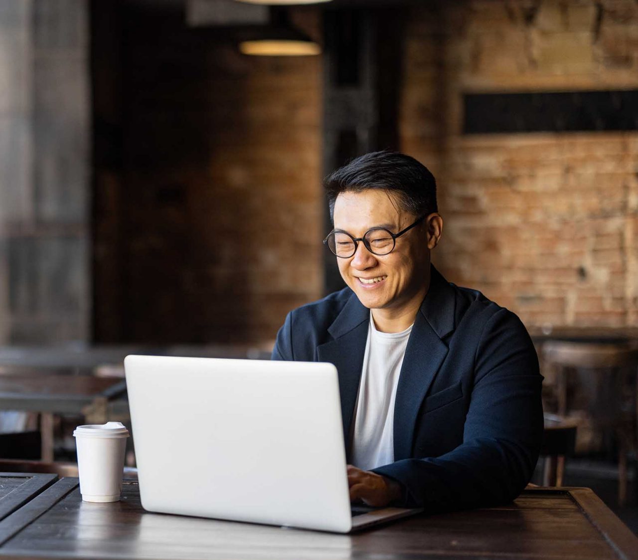 Asian businessman typing on laptop during work in cafe. Concept of remote and freelance work. Smiling adult successful man wearing suit and glasses sitting at wooden desk. Sunny day
